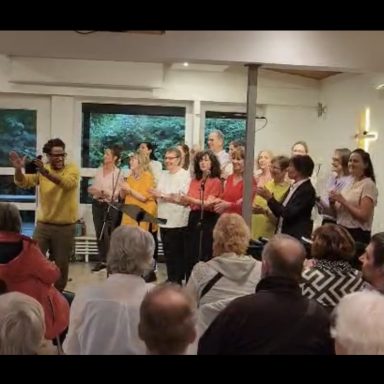 Der Chor steht auf der Bühne, hat Oberteile in fröhlichen Farben an und singt. Davor sieht man zahlreiche Besucher.