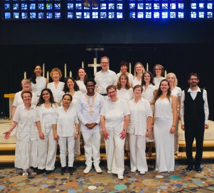 Der Chor steht vor dem Altar in der Kirche, alle sind ganz in Weiß gekleidet. Der Pianist, Christian Prüfling, steht daneben.