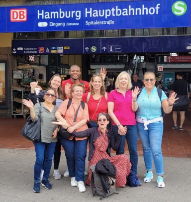 Ein Teil des ONE VOICE GOSPEL CHOIRS posiert unter dem Schild am Hamburger Hauptbahnhof. Alle sehen sehr fröhlich aus!