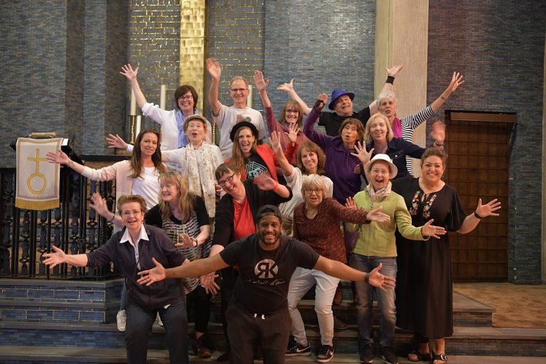 Foto des ONE VOICE GOSPEL CHOIR BERLIN vor dem Altar der Gustav-Adolf-Kirche Berlin. Alle recken freudig die Arme in die Höhe und winken.